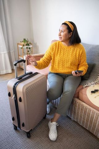 Mature woman preparing for trip at home pointing at luggage while holding smartphone