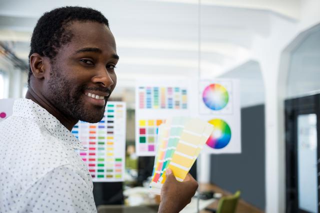 Male graphic designer holding a color swatch while smiling in a modern office. Ideal for use in articles or advertisements related to graphic design, creative professions, office environments, and color theory. Perfect for illustrating concepts of creativity, professional workspaces, and design projects.