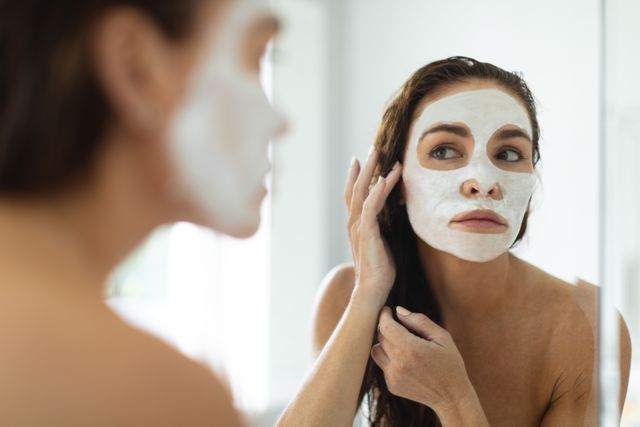 Adult woman applying a facial mask while looking into bathroom mirror, focusing on her self-care routine. Ideal for use in advertisements for beauty products, skincare blogs, wellness articles, and self-care promotion.