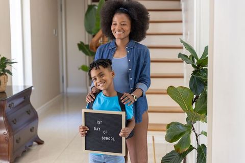 African-american mother supporting smiling son holding 1st day of school letterboard