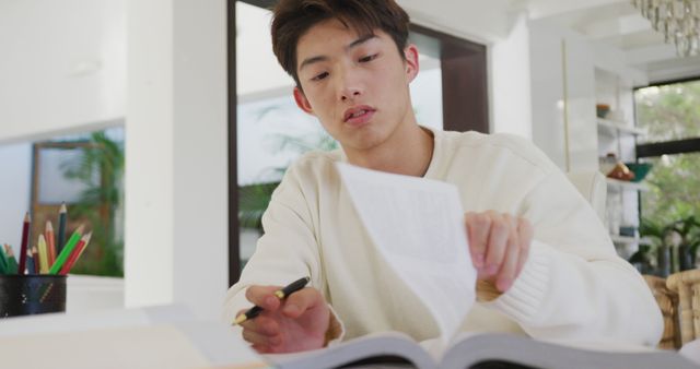 This stock photo captures an Asian male student studying at home with various books and notes spread out on the desk. He appears focused and intent on his work, which makes this image perfect for illustrating themes related to education, home studying, and the dedication of young students. This photo is ideal for educational content, blog posts on student life, remote learning, or promotional material for educational services.