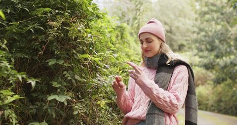 Woman in pink sweater and beanie exploring hedge and holding berry on woodland path