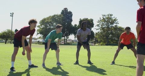 Diverse male teammates practicing fielding drill on sunny park lawn reacting to coach