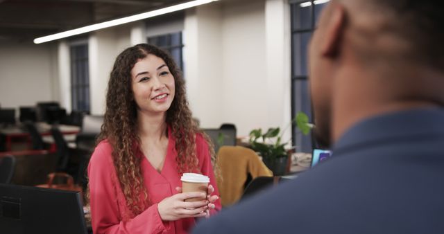 Two coworkers are engaging in a conversation in a modern office environment. One woman is holding a coffee cup, smiling while listening to the other person. The setting includes elements of a professional, open-plan office with desks, computers, and indoor plants. Useful for themes involving workplace culture, communication, collaboration, corporate environment, and daily office life.