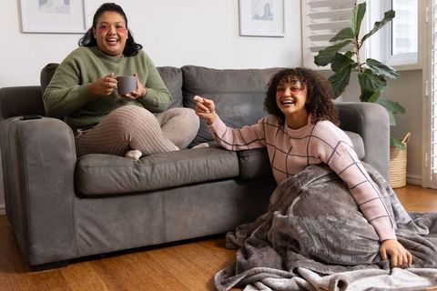 Laughing african american friends relaxing on sofa wrapped in blanket with coffee
