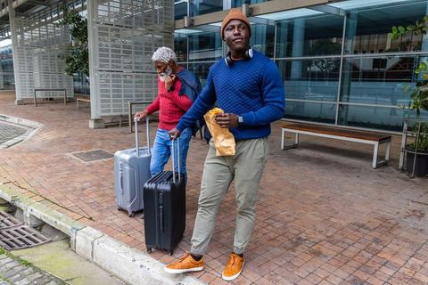 African american men walking with luggage and eating takeout outside modern glass terminal