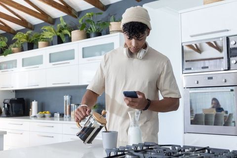 African american man pouring french press coffee while checking phone in modern kitchen