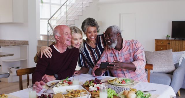 Seniors enjoying each other's company while sharing a meal, laughing, and using a smartphone to view photos or videos. Great for ads or content focused on senior lifestyle, technology adoption among the elderly, or multicultural friendships.