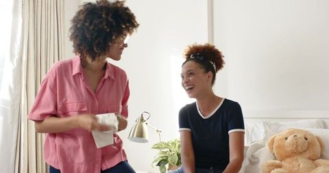 African American friends laughing and talking in cozy bedroom with teddy bear and lamp