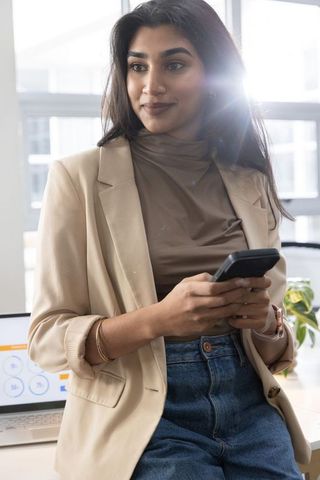 Indian businesswoman holding smartphone viewing laptop analytics with lens flare