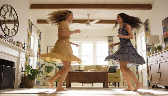 Friends twirling barefoot in sunlit living room wearing flowing dresses, joyful motion