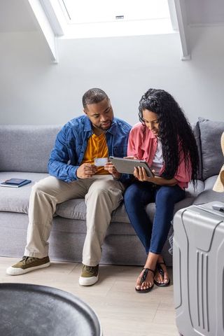 Couple planning trip on tablet while holding credit card beside suitcase in sunlit loft