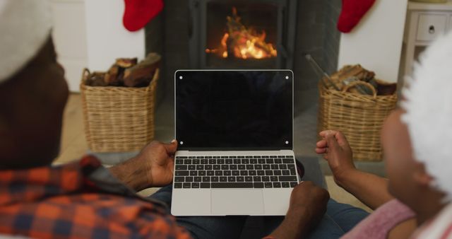 Couple sitting near fireplace using a laptop, enjoying warm and cozy ambiance of Christmas holidays. Perfect for depicting family bonding, festive season activities, or home comfort during winter. Can be used for holiday greetings, technology in everyday life, or promoting cozy home environments.