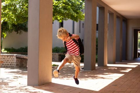 Young boy jumping through sunlit campus colonnade wearing backpack and striped shirt