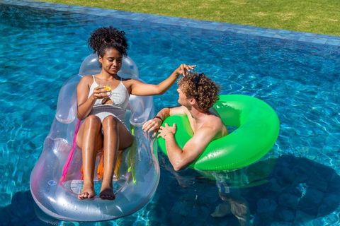 Diverse couple relaxing on clear lounge and green ring, sipping drink in backyard pool