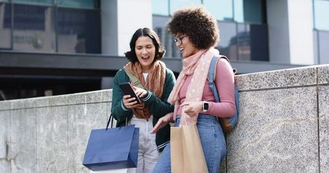 Two friends laughing and checking phone while shopping, leaning on stone wall in urban plaza