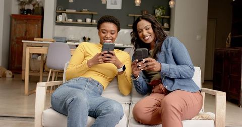 Two Female Friends Sitting on Sofa Using Smartphones Indoors