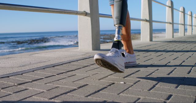 Person with a prosthetic leg walking along a coastal path, with the ocean in the background. The scene is filled with sunlight and conveys themes of mobility, rehabilitation, and an active lifestyle. Ideal for use in medical and health publications, promoting inspirational and motivational content, or illustrating themes of resilience and determination.