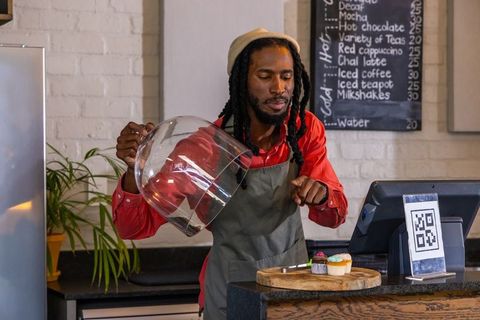 Barista lifting glass dome revealing cupcakes on counter with qr code and chalkboard menu