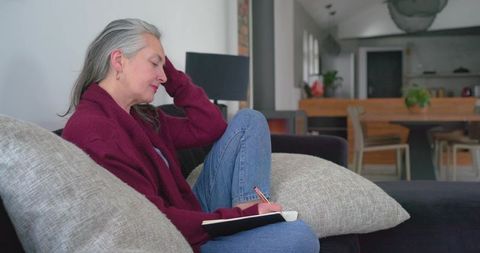 Mature woman journaling on sofa in cozy open-plan living room wearing burgundy sweater
