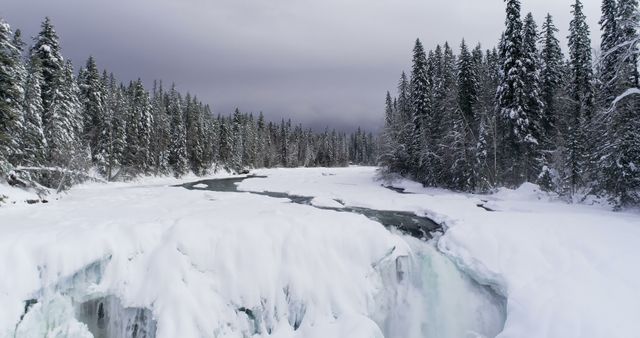 A stunning winter scene featuring a frozen river cutting through a snow-covered landscape lined with dense pine trees under a cloudy sky. Ideal for use in nature blogs, travel websites, winter-themed postcards, and seasonal promotional materials.