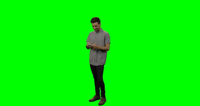 Image depicts a young man casually texting on a smartphone with a green screen background. It is ideal for technology, communication, and marketing uses. The green screen enables easy background manipulation for various promotional materials, advertisements, or presentations.