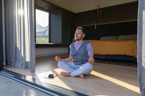 Mature asian man meditating cross-legged in sunlit minimalist bedroom near open sliding door