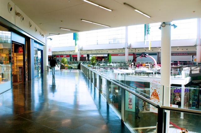Modern shopping mall with spacious design featuring bright natural lighting, glass railings, and various retail stores. Ideal for use in articles about retail industry trends, commercial real estate, shopping experiences, or architectural design.