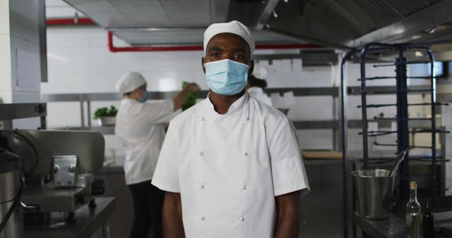 Portrait of african american male chef wearing face mask with arms crossed. Health and hygiene in restaurant kitchen during coronavirus covid 19 pandemic.