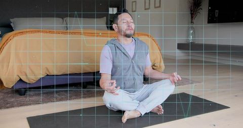 Meditating asian man on yoga mat in bedroom with digital grid overlay for wellness