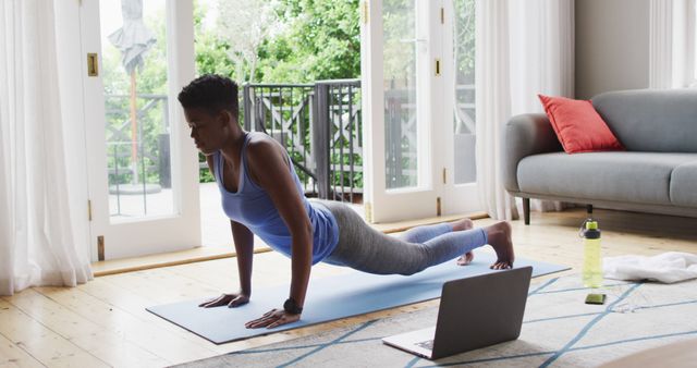 Woman performing yoga pose on mat in living room, following an online class on laptop. Bright natural light from large windows creates serene atmosphere. Suitable for fitness blogs, remote workout programs, and healthy lifestyle promotions.