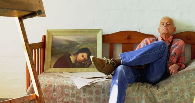 Elderly man in casual clothing relaxing in a home art studio, sitting on a wooden bench beside his artwork. Suitable for themes involving artistic environments, leisure, creativity in old age, and peaceful moments.