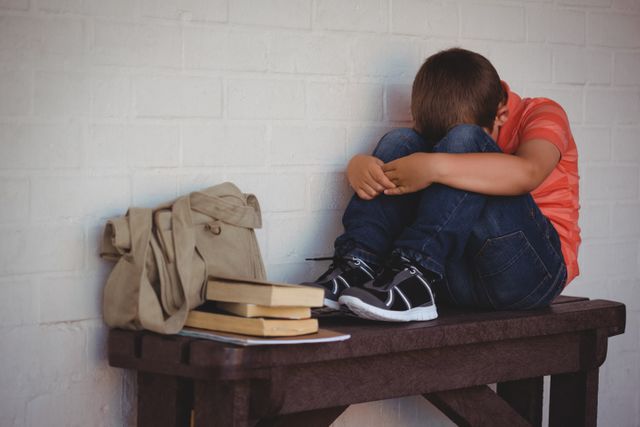 Young boy sitting on a bench in a school corridor, looking sad and lonely. His school bag and books are placed beside him. This image can be used to depict themes of bullying, childhood emotional distress, mental health issues in students, and the challenges faced by young students in educational environments.