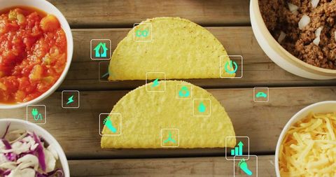 Two hard corn taco shells sitting on wooden plank with sustainability icons and ingredients