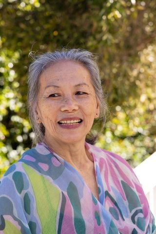 Senior asian woman smiling on sunlit porch wearing colorful pastel shawl, peaceful outdoor portrait