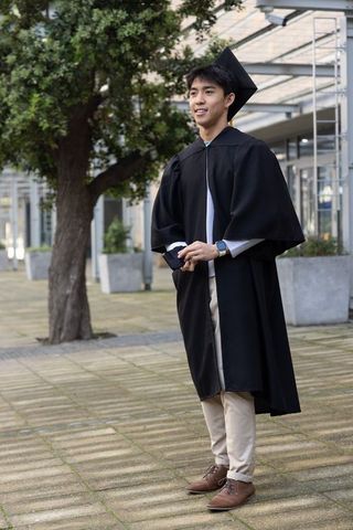 Asian male graduate standing on modern university campus wearing gown and mortarboard