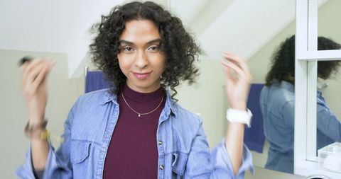 Non-binary person adjusting curly hair at bathroom mirror wearing denim shirt, necklaces