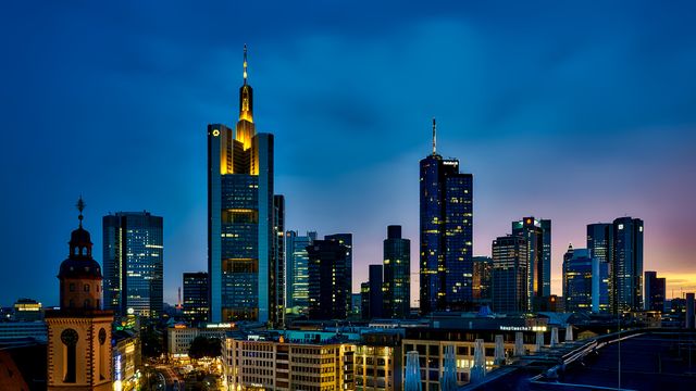 Captures the Frankfurt skyline at dusk with illuminated skyscrapers standing tall against a darkening sky. The vibrant urban landscape highlights the city's modern architecture and financial district. Perfect for content related to travel, urban development, business hubs, tourism, and architecture features.