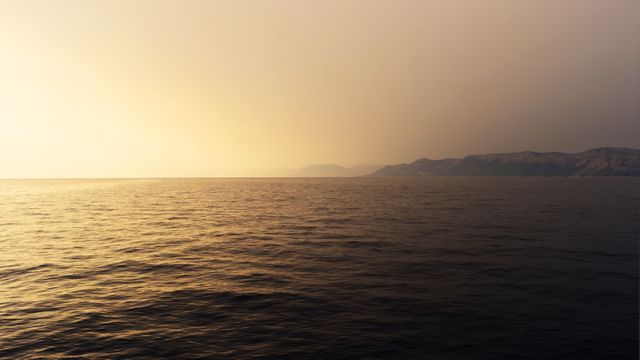Ocean calm at sunset, sun casting golden light across water. Distant mountains creating tranquil scene. Suitable for nature-themed designs, desktop backgrounds, travel articles, meditative content