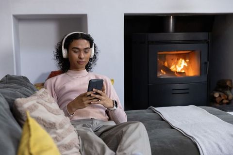 Non-binary person relaxing on sofa wearing headphones and using smartphone by fireplace
