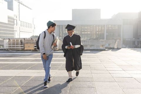 Happy graduate walking campus terrace with friend holding diploma and checking phone