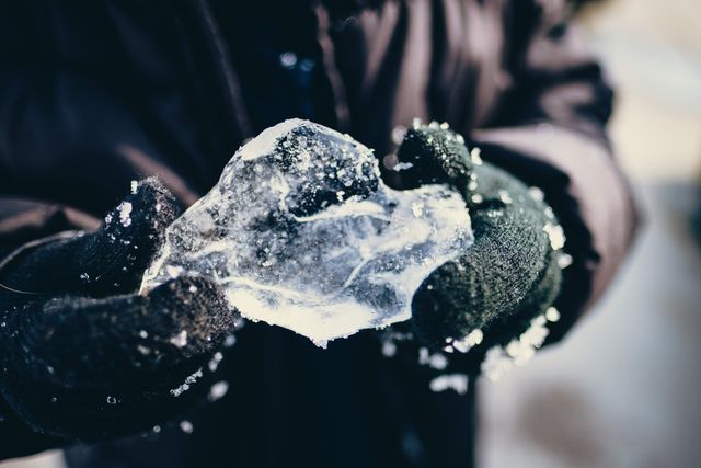 Close-up image shows gloved hands holding a translucent ice crystal outdoors during a cold winter day. This image can be used for illustrating themes related to winter, cold, nature, and activities in snowy environments. It is suitable for blog posts, advertisements about winter gear, and informational articles about natural ice formations.
