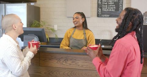 Smiling indian barista chatting with diverse customers at coffee counter holding red mugs
