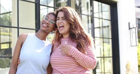 Two diverse women laughing outdoors by modern black-framed glass doors on sunny patio