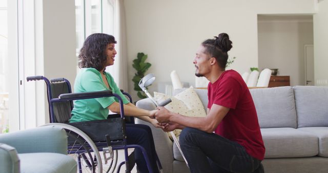 Happy biracial woman in wheelchair and smiling male partner sitting holding hands in living room. wellbeing and domestic lifestyle with physical disability.