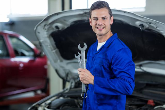 Smiling mechanic in blue uniform holding wrenches in an auto repair garage. Ideal for use in automotive service advertisements, repair shop promotions, and articles about car maintenance and professional mechanics.