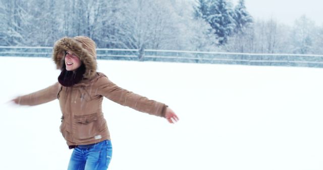 Woman standing outside in a snowy setting wearing a brown winter jacket with fur-lined hood and blue jeans, spreading arms joyfully. Ideal for promotions related to winter activities, outerwear fashion, or nature-related themes.