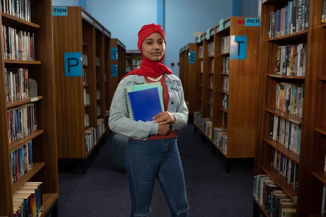 Front view of an Asian female student wearing a red hijab and jeans studying in a library, holding a book in hands.