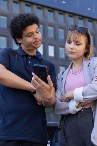 Young diverse peers checking smartphone near modern office facade, teamwork collaboration