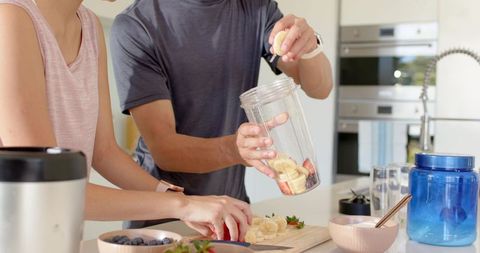 Diverse couple preparing fruit smoothie in modern kitchen, slicing bananas and berries